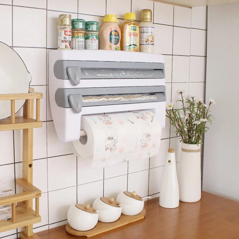 A wall-mounted 3-in-1 foil cutter and kitchen roll holder in white color, with a sliding cutter mechanism and a detachable rod for hanging paper towels or cloths, displayed in a kitchen setting with various items on the shelf below.