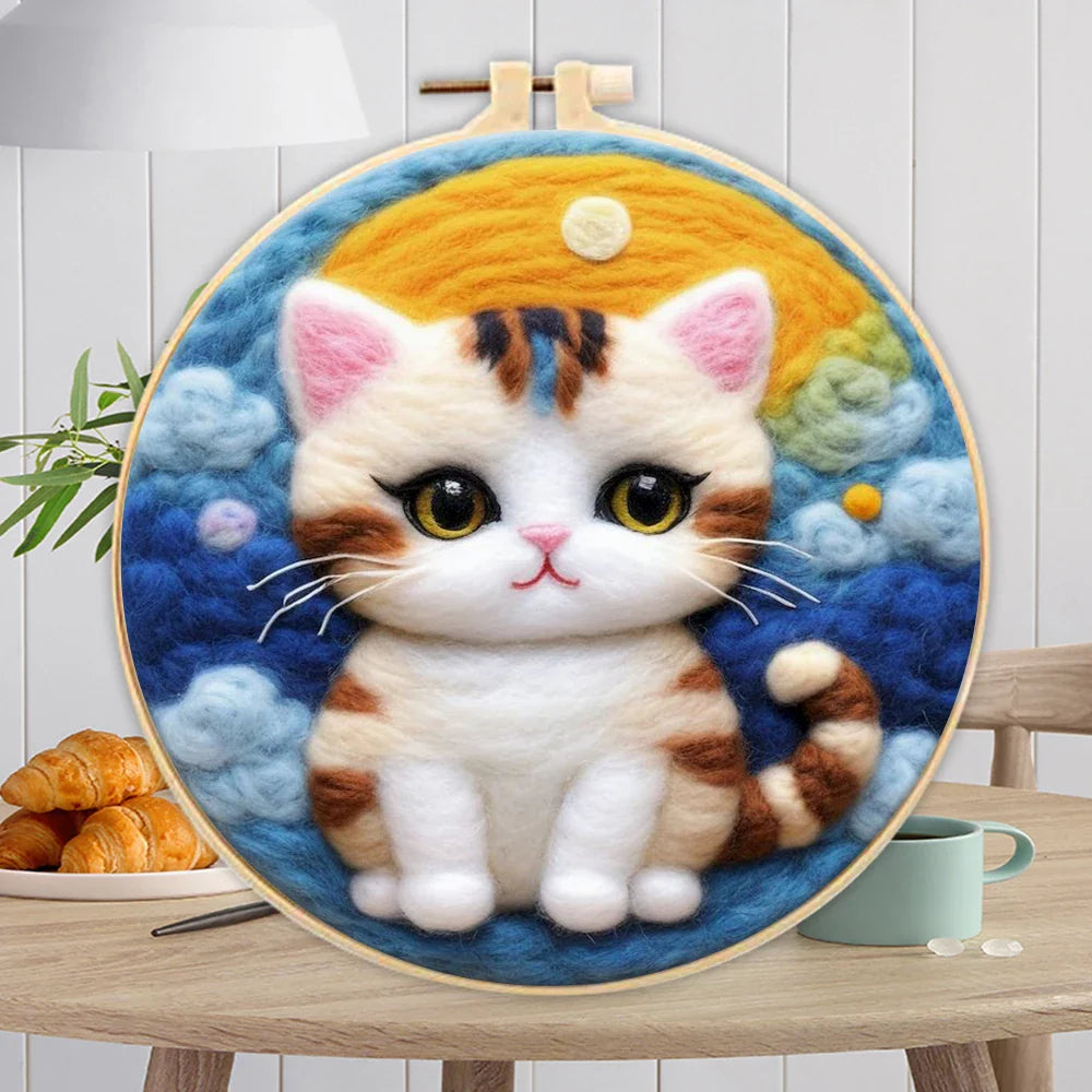 A person's hands holding a DIY felting kit with a pre-printed felt cloth depicting a white cat with orange spots, surrounded by colorful wool in the background.