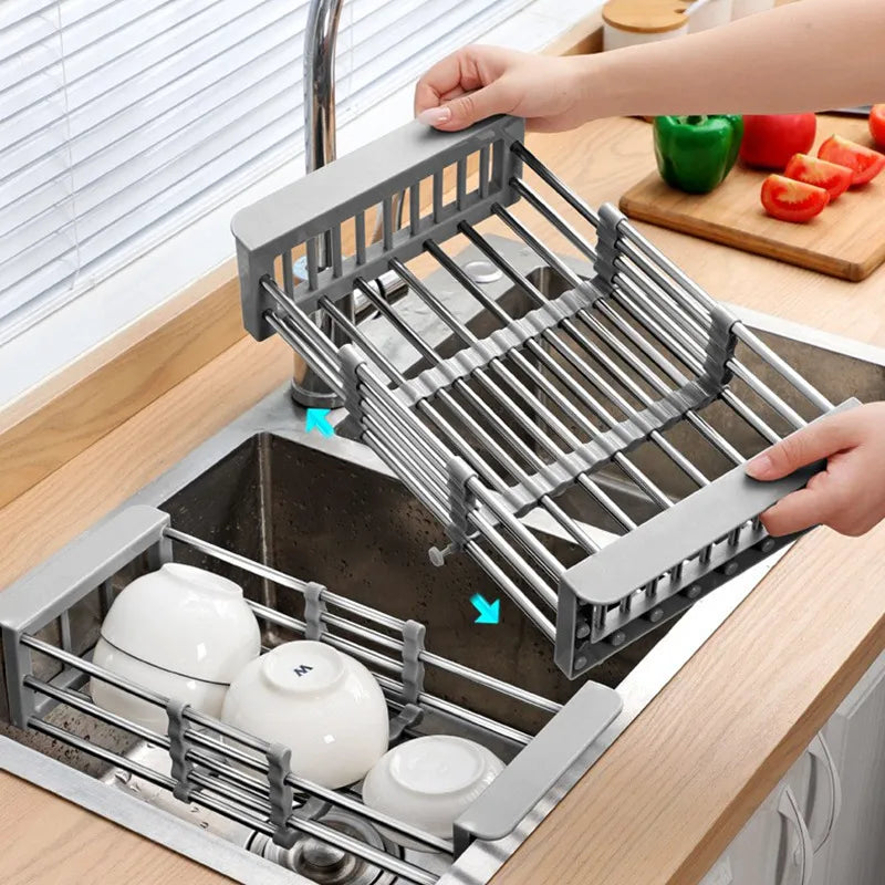 A stainless steel sink basket with telescopic rods being adjusted to fit inside a sink, with dishes and vegetables placed on it.