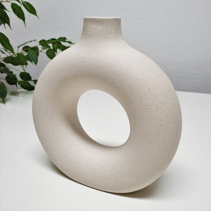 Two round ceramic hole vases with different sizes, one smaller and one larger, placed on a surface with dried plants inside the smaller one, with a painting and a wooden chair in the background.