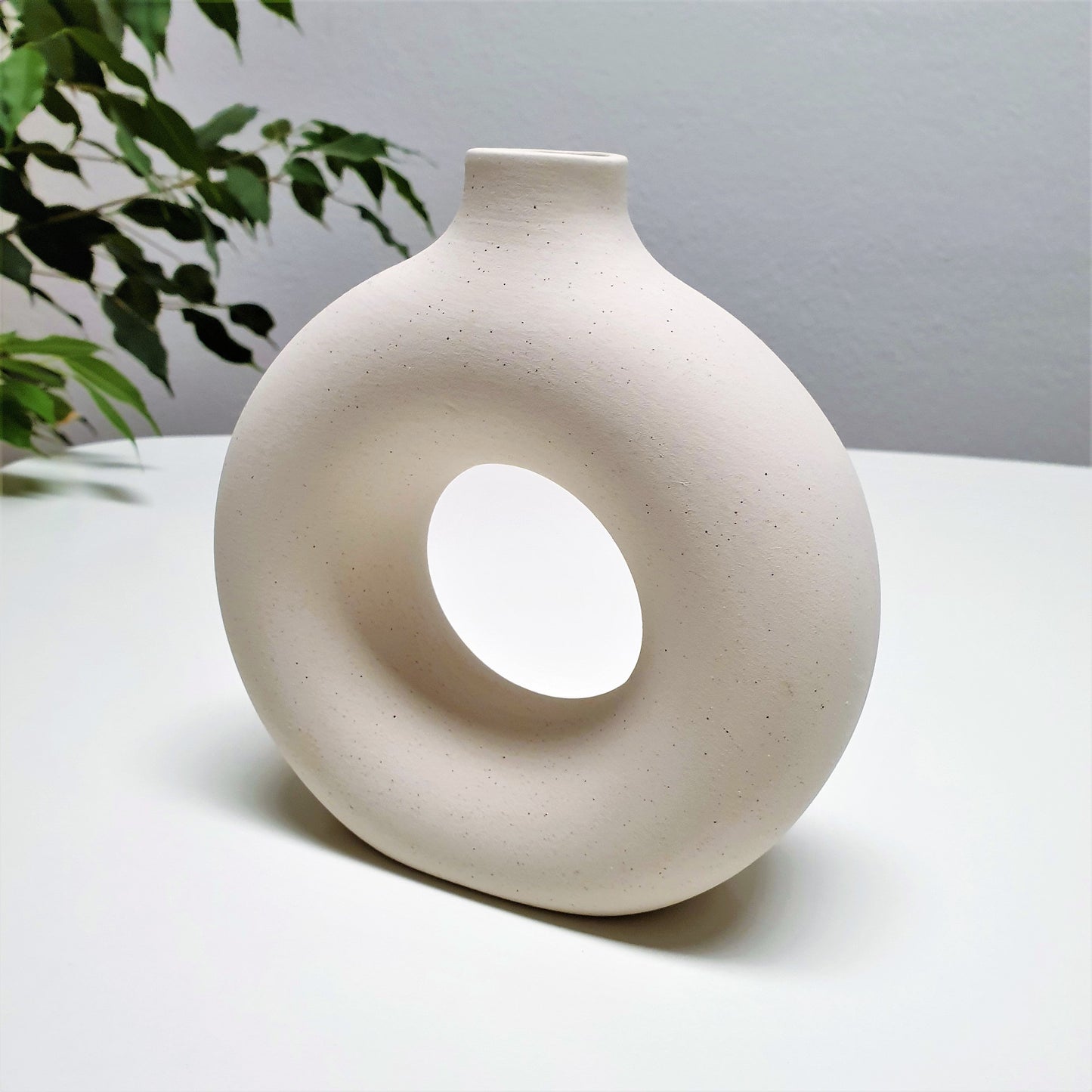 Two round ceramic hole vases with different sizes, one smaller and one larger, placed on a surface with dried plants inside the smaller one, with a painting and a wooden chair in the background.