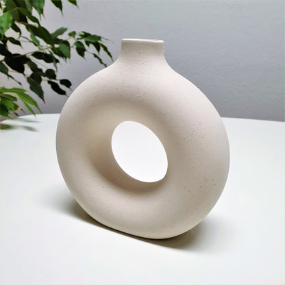 Two round ceramic hole vases with different sizes, one smaller and one larger, placed on a surface with dried plants inside the smaller one, with a painting and a wooden chair in the background.