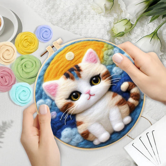 A person's hands holding a DIY felting kit with a pre-printed felt cloth depicting a white cat with orange spots, surrounded by colorful wool in the background.
