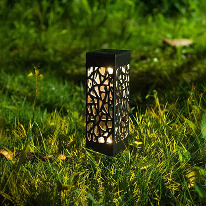 A solar garden path light with a rectangular black frame and intricate cut-out pattern, illuminated and placed on grass.